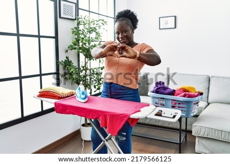 Young african woman ironing clothes at home smiling in love doing heart symbol shape with hands. romantic concept. 