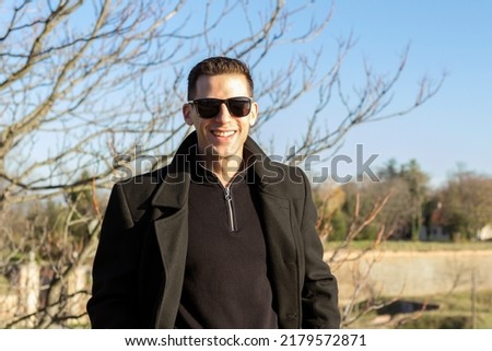 Stock photo of a good looking man, casually dressed in black, posing and smiling at the camera during an autumn day.