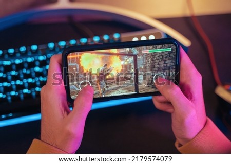 Close up of male pro gamer's hands holding smartphone with online video shooter game. Cyber sportsman playing mobile games on his cellphone. Professional keyboard on background. Top view. Neon lights