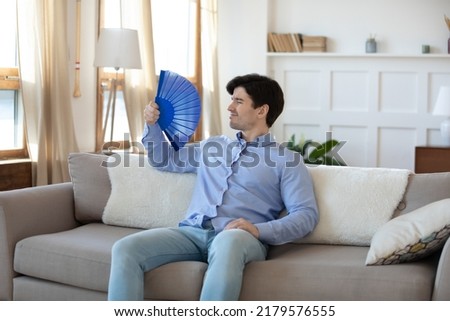Overheated young man owner renter tenant of house flat resting on sofa in living room saving himself from heat using paper weaver understanding problem of bad ventilation and lack of air conditioning. Royalty-Free Stock Photo #2179576555