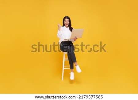 Young woman asian happy smiling. While her using laptop sitting on white chair and looking isolate on yellow background.