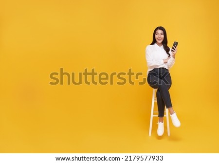 Happy young Asian woman showing mobile phone. While her sitting on white chair and looking on yellow copy space background.