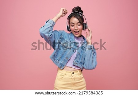 Happy beautiful Asian teen woman in yellow glasses listening music in headphones and dance on pink background. Royalty-Free Stock Photo #2179584365
