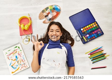 Young brunette woman lying on the floor at art studio around artist things doing ok sign with fingers, smiling friendly gesturing excellent symbol 