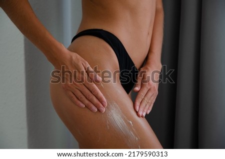 A close-up picture of a young slim tanned woman spreading white cream on her hips with her hands. The picture shows body parts of a young woman moisturising her hips.