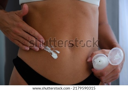 A close-up photo of a slim belly of a young woman and her hands spreading white cream on her tummy. She is wearing black and white underwear.
