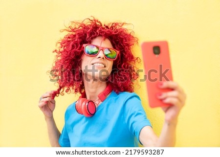 portrait of a smiling young woman with red afro hair wearing a blue t-shirt and sunglasses, taking a selfie photo touching her hair on yellow background, red headphones around her neck