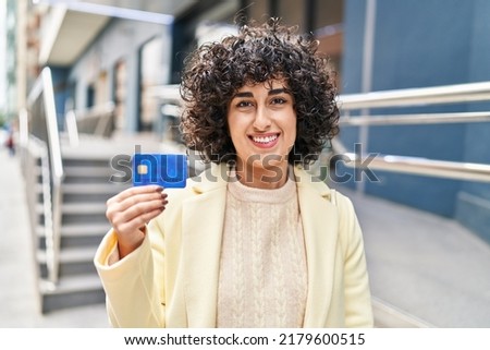 Young brunette woman with curly hair holding credit card looking positive and happy standing and smiling with a confident smile showing teeth 