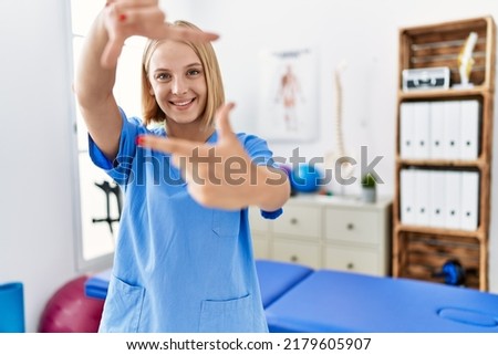 Young caucasian physiotherapist woman working at pain recovery clinic smiling making frame with hands and fingers with happy face. creativity and photography concept. 