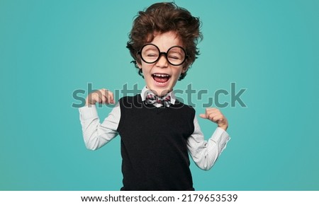 Cute little nerd in school uniform and glasses with big head laughing and showing biceps during studies against turquoise background