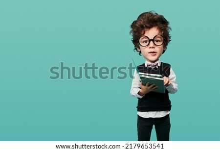 Adorable funny little boy with wavy dark hair and big head in round eyeglasses and classy outfit, holding books and looking at camera against turquoise background