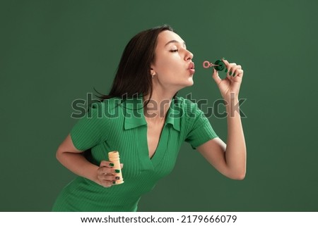 Portrait of beautiful young girl posing, blowing soap bubbles isolated over green studio background. Summertime fun. Concept of emotions, facial expression, lifestyle, fashion, youth culture Royalty-Free Stock Photo #2179666079