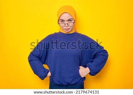 Annoyed young Asian Muslim woman dressed in casual sweater holding hands on waist and looking at camera isolated over yellow background