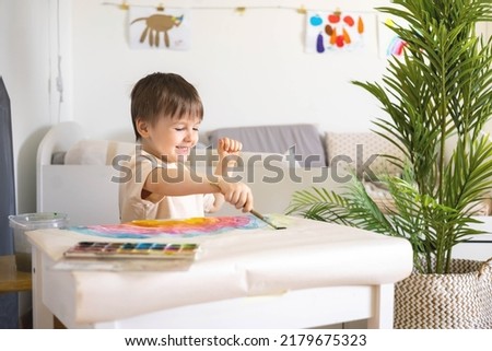 Confident cute male kid art drawing multicolored rainbow arch use paints and brush on table at kindergarten home interior. Focused baby boy enjoying artwork early development creative activity