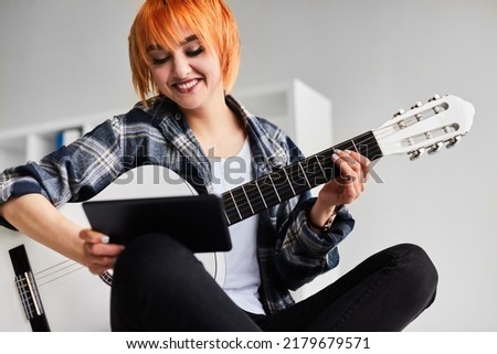 Crop positive young female musician with short dyed red hair and bright makeup in checkered shirt, smiling and watching tutorial video on tablet while playing acoustic guitar in white room