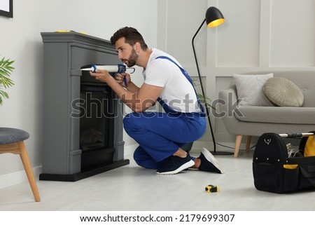 Professional technician sealing electric fireplace with caulk near white wall in room Royalty-Free Stock Photo #2179699307