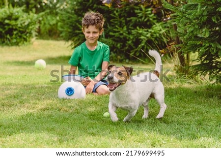 Happy boy and dog playing with automatic throw and fetch machine launching tennis balls Royalty-Free Stock Photo #2179699495