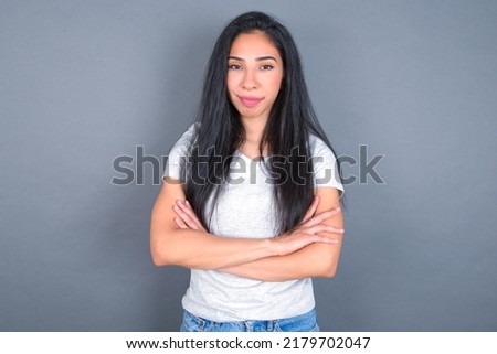 Self confident serious calm young beautiful brunette woman wearing white T-shirt over white wall stands with arms folded. Shows professional vibe stands in assertive pose.