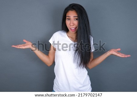 So what? Portrait of arrogant young beautiful brunette woman wearing grey T-shirt over white wall shrugging hands sideways smiling gasping indifferent, telling something obvious. Royalty-Free Stock Photo #2179702147