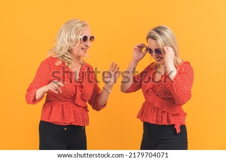 Two european blonde millenial women in red dotted shirts and sunglasses dancing, singing and having fun. Studio shot on orange background. High quality photo