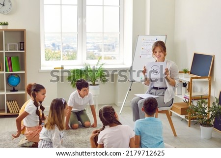 We learn to talk about emotions. Friendly female school psychologist talks about emotions during meeting with group of elementary school students. Woman shows smiling emoticon to students.