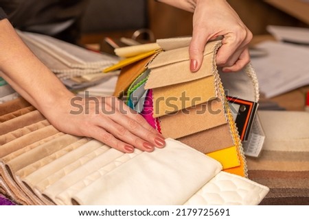 Fabric swatches in different colors are stacked for selection. A woman chooses upholstery colors for furniture and interiors. Touches the texture of the fabric, hands on the material close-up. Royalty-Free Stock Photo #2179725691