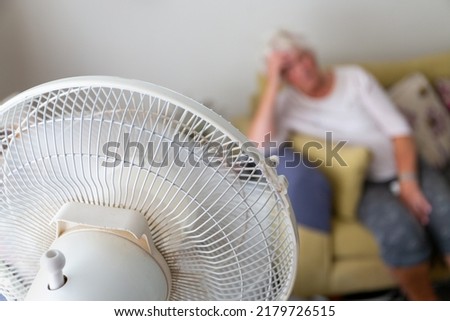 Senior woman on a sofa suffering from excessive heat during a heatwave and being cooled by an oscillating electrical fan in the foreground Royalty-Free Stock Photo #2179726515