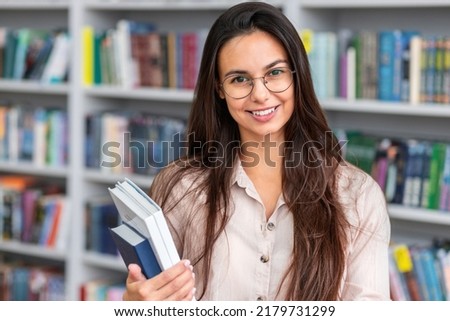 Portrait of beautiful university student woman with eye glasses standing with books against the background of bookshelves, in campus library looking at the camera and smiling