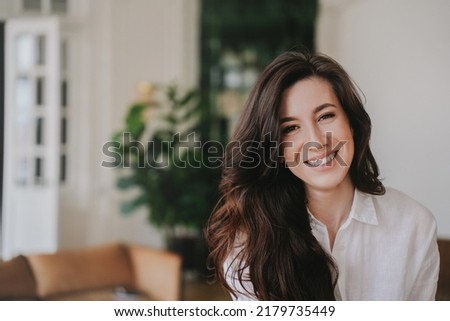 Portrait of hispanic brunette woman with wavy dark hair wearing white shirt, looking at camera, smiling at home. Successful entrepreneur toothy smiling, happy to develop business using new strategies. Royalty-Free Stock Photo #2179735449