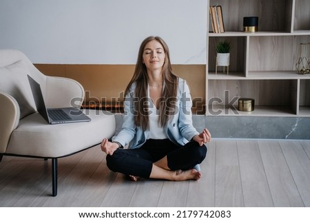 Emotionally balanced young caucasian woman in blue  sitting on floor at chair with laptop eyes closed in meditative pose, relaxing at home at break of remote working, against fireplace. Businesswoman