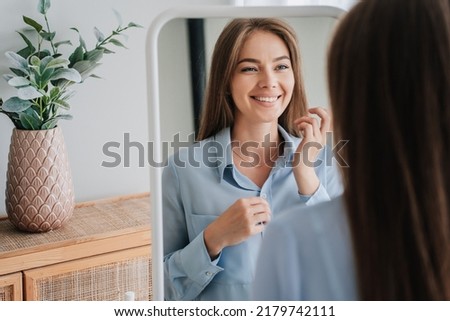 Cheerful young European woman in blue shirt standing at mirror toothy smiling satisfied by her skin health. Successful caucasian blonde woman toothy smiling at home, enjoying sunny morning at bedroom. Royalty-Free Stock Photo #2179742111
