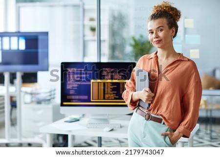 Portrait of young female programmer with digital tablet looking at camera while standing at her workplace with computer Royalty-Free Stock Photo #2179743047