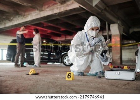 Young criminological expert in coveralls inspecting empty bottle on crime scene while squatting in front of open briefcase with working supplies