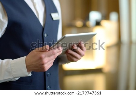 Digital tablet in hands of young male receptionist of luxurious modern hotel standing in lounge by entrance and meeting new guests Royalty-Free Stock Photo #2179747361
