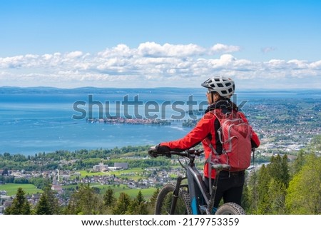 happy, active senior woman cycling above Lake Constance, Bodensee and  admiring the awesome view over Lake Constance to city of Lindau in background