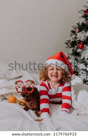 little girl in striped pajamas and a Santa hat and dog in funny glasses with Santa Claus are lying in bed on a white sheet against the background of Christmas tree. space for text. High quality photo