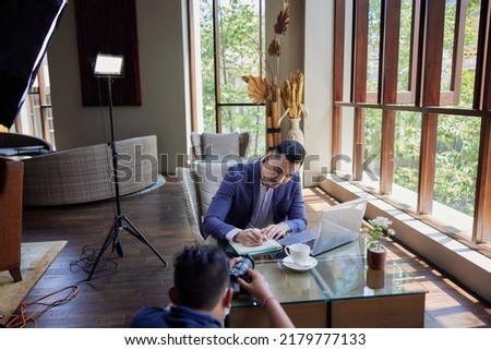 Professional male photographer takings pictures of businessman sitting with laptop writing notes. Behind the scene in photography industry. Filming stock photography content