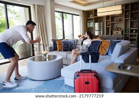 Husband taking photo of wife using smartphone in hotel room after check-in. Happy young asian couple with suitcases having fun in living room of luxury villa