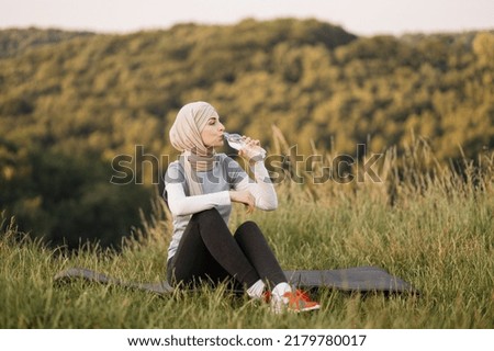 Attractive muslim woman in hijab resting while sitting on the mat and drinking water from a bottle outdoors in park . Healthy lifestyle concept.