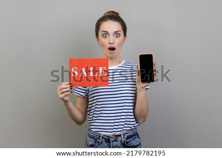 Portrait of shocked surprised woman wearing striped T-shirt standing with open mouth, holding sale card and cell phone with empty display. Indoor studio shot isolated on gray background.