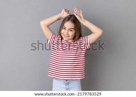 Portrait of little girl wearing striped T-shirt showing bunny ears gesture, holding hands on head and smiling at camera, having fun. Indoor studio shot isolated on gray background.