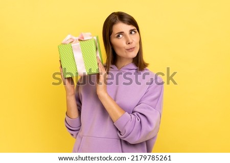 Portrait of curious dark haired woman holding shaking green wrapped present box, thinks what is inside, wearing purple hoodie. Indoor studio shot isolated on yellow background.