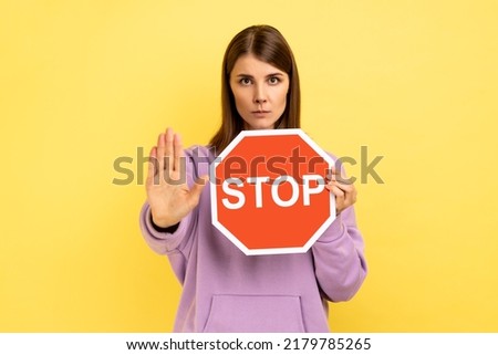 Portrait of strict bossy woman showing stop gesture and holding red stop road sign, prohibitions and restrictions, wearing purple hoodie. Indoor studio shot isolated on yellow background.