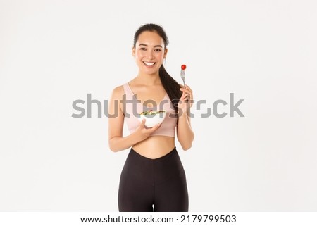Sport, wellbeing and active lifestyle concept. Smiling healthy and slim asian girl in fitness clothing, holding salad and pick tomato on fork, staying fit with diet and special workout Royalty-Free Stock Photo #2179799503