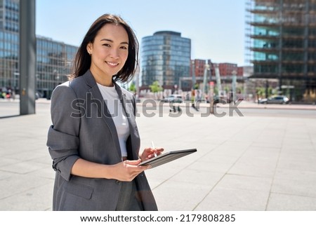 Young happy Asian business woman entrepreneur wearing suit holding digital tablet standing in big city on busy street, using smart business software tech for online work on pad computer outdoor. Royalty-Free Stock Photo #2179808285