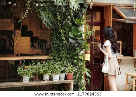 Young woman opening door of modern cozy coffeeshop with walls decorated with lush plants Royalty-Free Stock Photo #2179824563