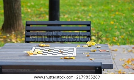 A serene picture of an empty metallic chess table and grey structured wooden bench waiting for the game in an autumn park full of fallen yellow leaves. High quality photography.