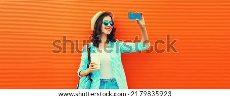 Portrait of happy smiling young woman taking selfie with smartphone with cup of coffee wearing summer straw hat, backpack on orange background, blank copy space for advertising text