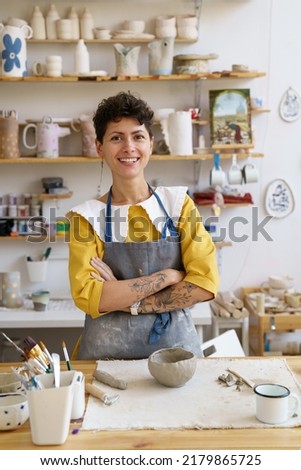 Young ceramist small business and pottery studio owner in workshop. Female craft crockery and potter tableware master at workplace wearing apron happy smiling. Ceramics retail store entrepreneurship