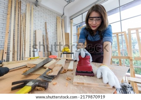 Ethnic woman in apron and goggles polishing lumber board on table while working in professional joinery